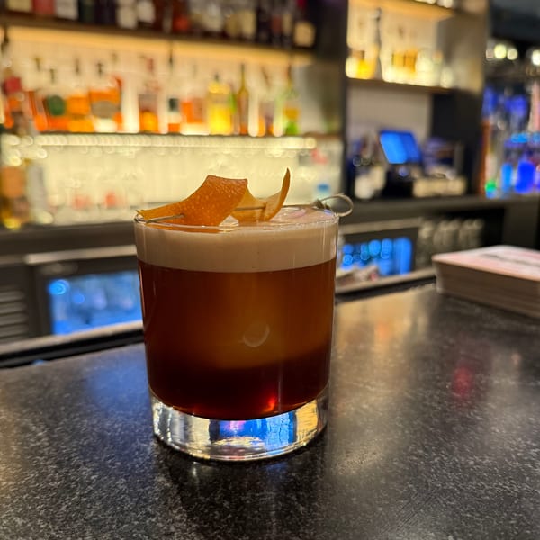 A dark cocktail with a foamy top and orange peel garnish sits on a bar counter.
