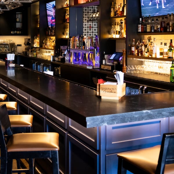 A long, dark bar counter with golden bar stools, liquor bottles, beer taps, and TVs.