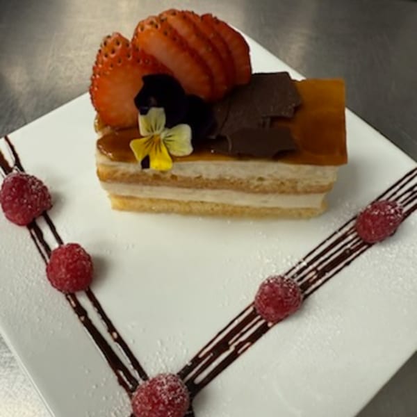Layered dessert with strawberries, chocolate, and an edible flower, garnished with raspberries and chocolate drizzle.