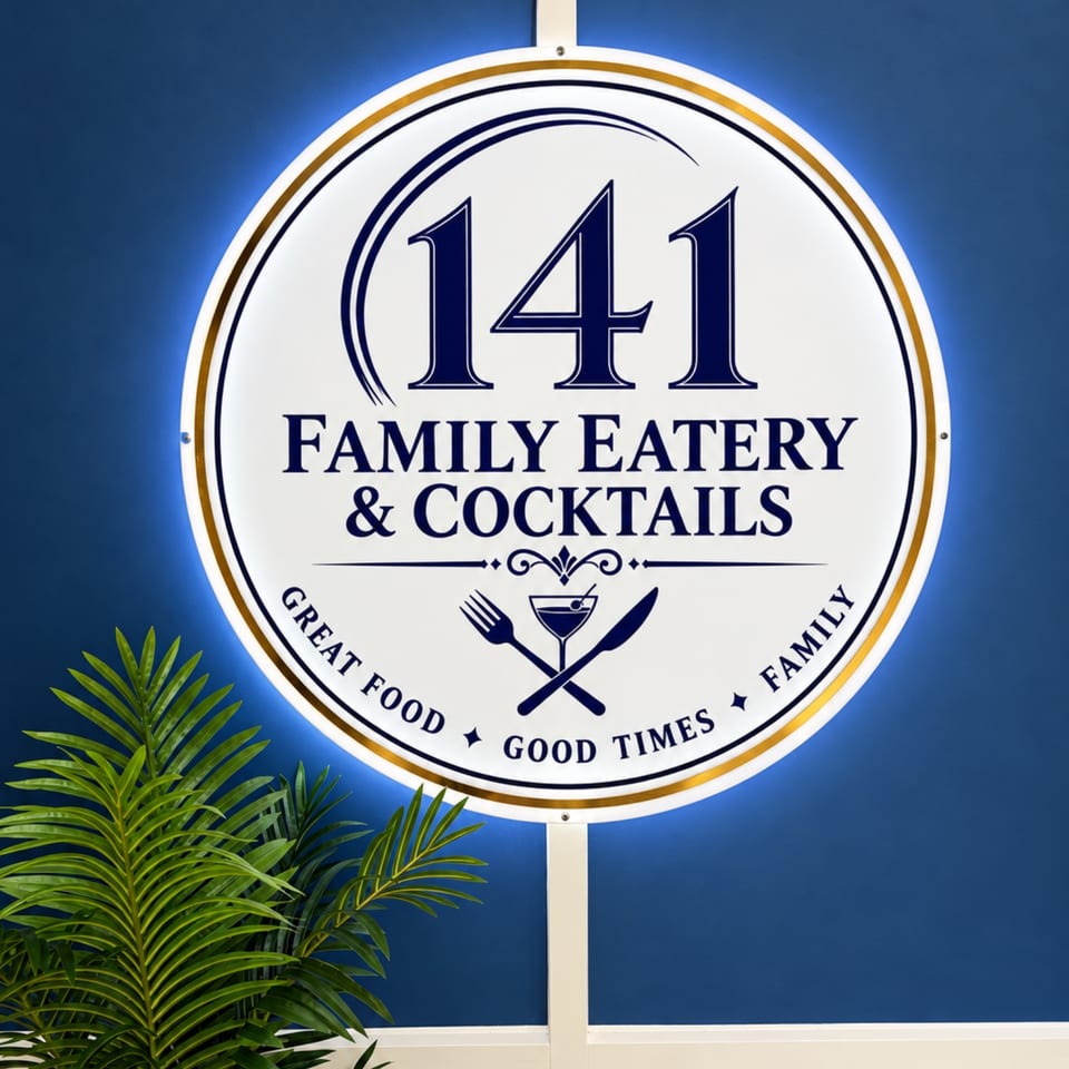 Illuminated circular sign for 141 Family Eatery & Cocktails against a blue wall, with tropical plants below.