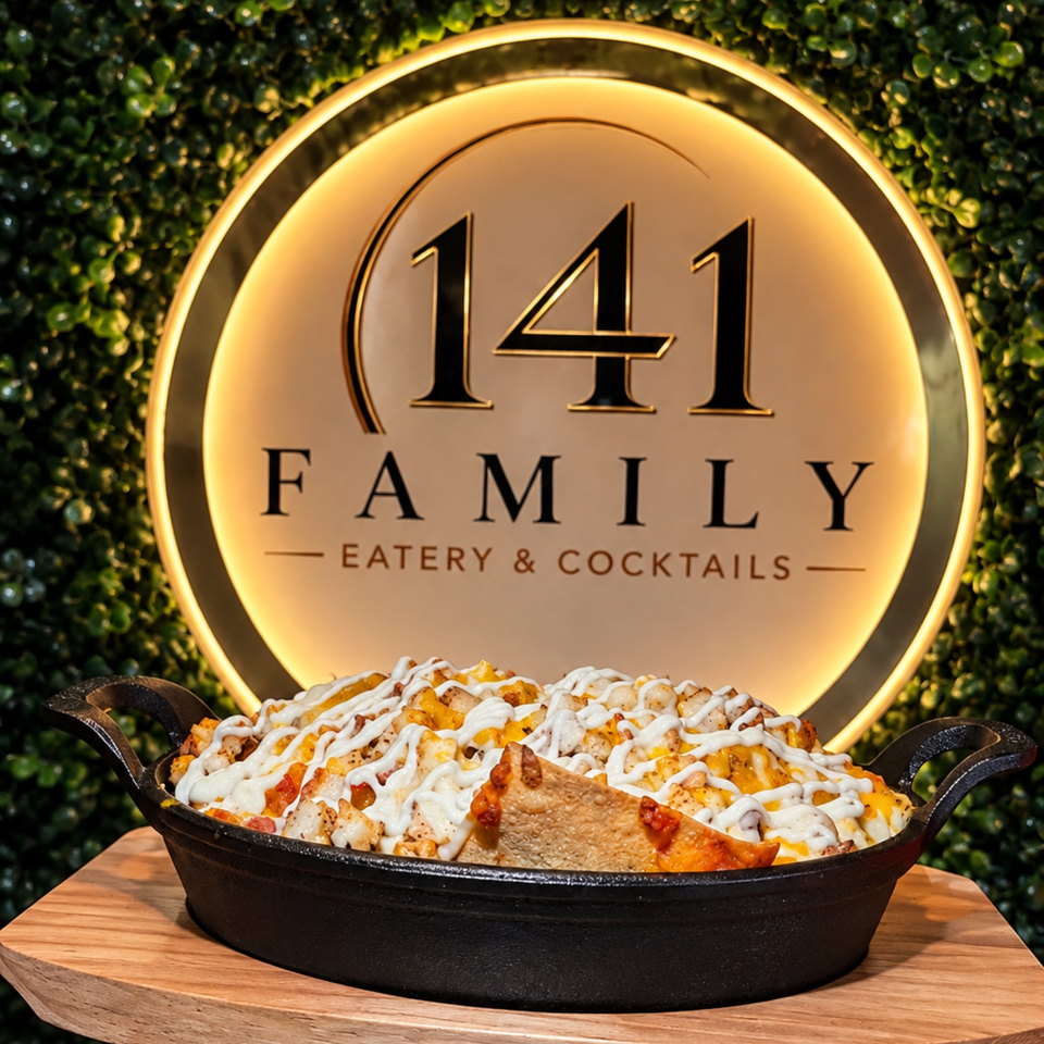 Loaded skillet of cheesy fries drizzled with white sauce, displayed before the glowing 141 Family Eatery & Cocktails sign.
