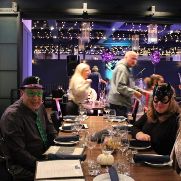 People in costumes, including Riddler and Catwoman, seated at a Bar 141 dining table with string lights.