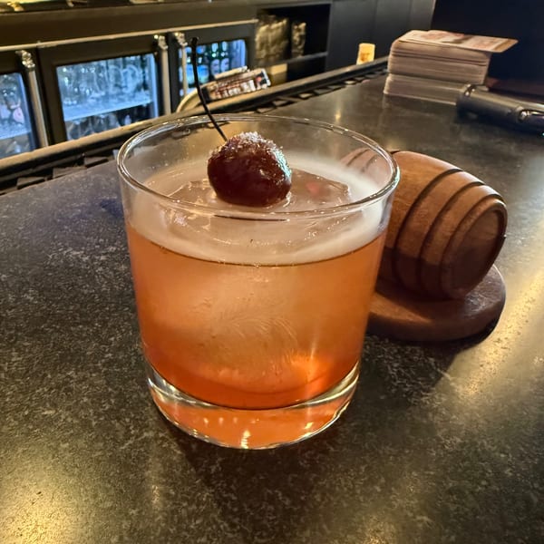 An orange cocktail with a large ice cube and a sugared cherry on a dark bar counter.