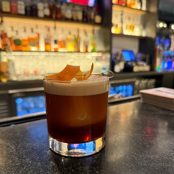 A cocktail with foamy top and orange peel garnish sits on a dark bar counter with bottles in the background.