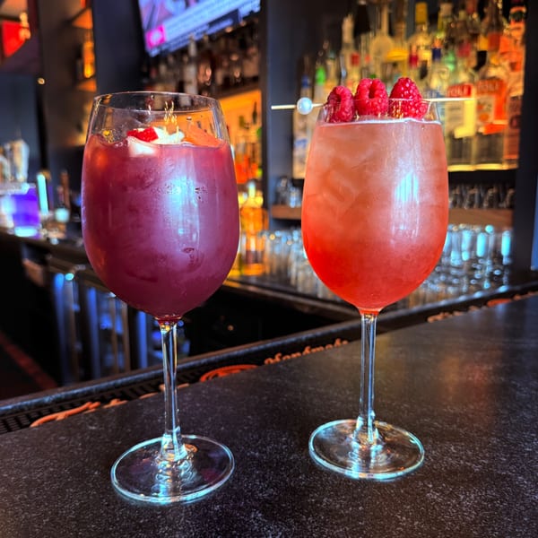 Two vibrant cocktails, one red with fruit and one pink with raspberries, sit on a dark bar.