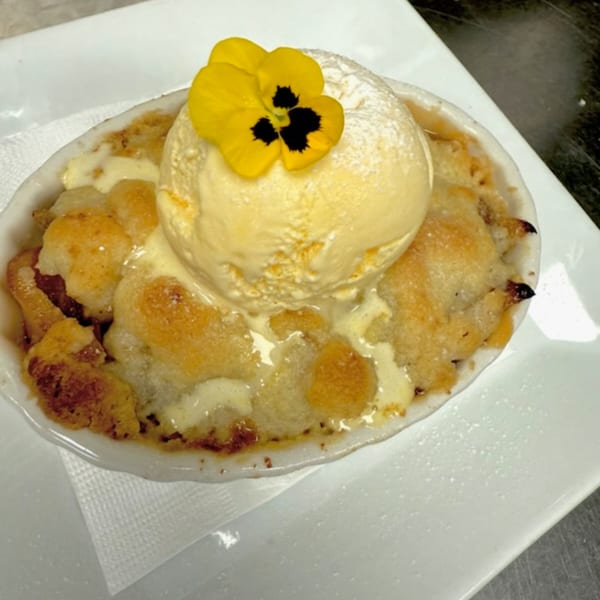 Warm fruit crumble topped with a scoop of vanilla ice cream and a yellow edible flower.
