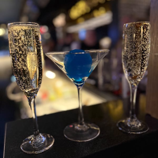Two champagne flutes and a martini glass with a blue ice sphere, on a dark surface.