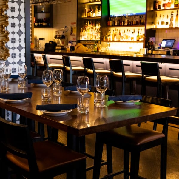 A long wooden table set with plates, glasses, and blue napkins, facing a well-stocked bar.