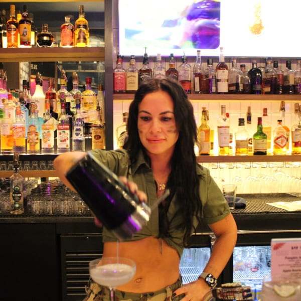 A Bar 141 bartender pours a cocktail from a shaker into a glass behind a well-stocked bar.