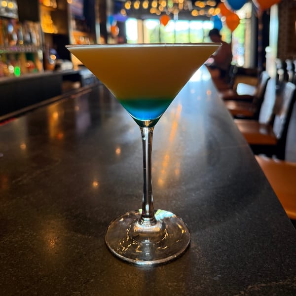 A martini glass with a yellow and blue layered drink sits on a dark bar counter.