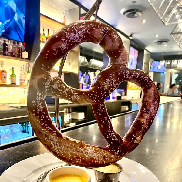 A large, salted pretzel hangs from a stand with two dipping sauces on a bar counter.